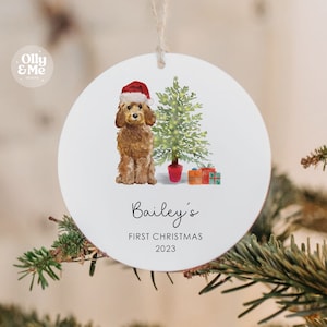 May include: A round Christmas ornament featuring a brown dog wearing a Santa hat, a Christmas tree, and wrapped gifts. The text "Bailey's FIRST CHRISTMAS 2023" is printed below the dog. The ornament is hanging from a string.