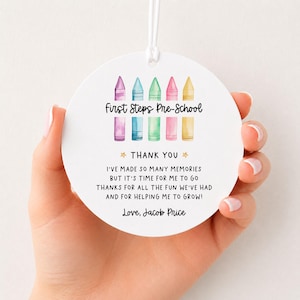 May include: A round white ornament with a white string featuring a watercolour illustration of five colourful crayons and the text First Steps Pre-School, Thank You, I've made so many memories but it's time for me to go thanks for all the fun we've had and for helping me to grow! Love, Jacob Price.