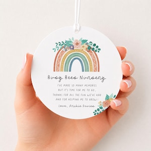 May include: A round white ceramic ornament featuring a colorful rainbow illustration topped with flowers. The text reads: Busy Bees Nursery, I've made so many memories but it's time for me to go, thanks for all the fun we've had and for helping me to grow! Love, Archie Davies.