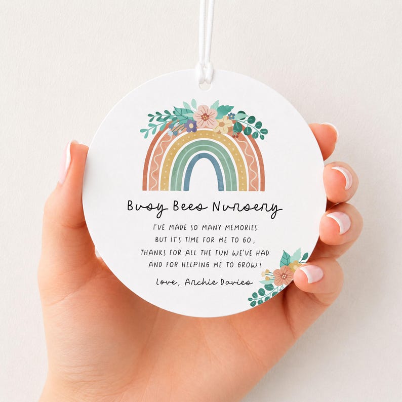 May include: A round white ceramic ornament featuring a colorful rainbow illustration topped with flowers. The text reads: Busy Bees Nursery, I've made so many memories but it's time for me to go, thanks for all the fun we've had and for helping me to grow! Love, Archie Davies.