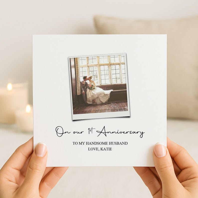 May include: A square white greeting card featuring a central polaroid-style photograph of a couple in wedding attire sitting by a window. Below the photo, black text reads: On our 1st Anniversary, TO MY HANDSOME HUSBAND, LOVE, KATIE.