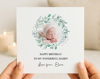 May include: A square white greeting card featuring a watercolor wreath of green leaves and blue berries surrounding a photograph of a baby wearing a white knit hat. The text reads: HAPPY BIRTHDAY TO MY WONDERFUL DADDY. Love from. Elias.