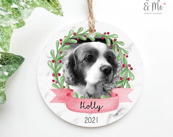 May include: A white round Christmas ornament with a black and white photo of a dog inside a green wreath with red berries. The wreath has a pink ribbon with the word "Holly" and the year "2021" printed below.