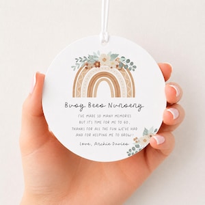 May include: A round white ceramic ornament featuring a printed illustration of a tan and brown rainbow topped with flowers and eucalyptus leaves. The text reads Busy Bees Nursery, followed by a thank you message and the name Archie Davies.