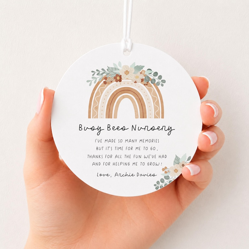 May include: A round white ceramic ornament featuring a printed illustration of a tan and brown rainbow topped with flowers and eucalyptus leaves. The text reads Busy Bees Nursery, followed by a thank you message and the name Archie Davies.