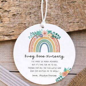 May include: A round white ornament featuring a colorful rainbow graphic topped with flowers. The text reads: Busy Bees Nursery, I've made so many memories but it's time for me to go, thanks for all the fun we've had and for helping me to grow! Love, Archie Davies. It includes a white string.