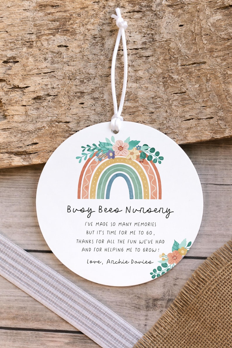 May include: A round white ornament featuring a colorful rainbow graphic topped with flowers. The text reads: Busy Bees Nursery, I've made so many memories but it's time for me to go, thanks for all the fun we've had and for helping me to grow! Love, Archie Davies. It includes a white string.