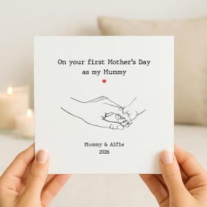 Op de afbeelding: Een vierkante witte wenskaart met een zwarte lijntekening van een volwassen hand die een babyhandje vasthoudt. De tekst luidt: On your first Mother's Day as my Mummy, met een klein rood hartje. Onderaan staat: Mummy & Alfie 2026.