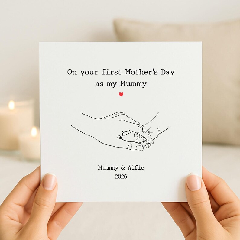 Op de afbeelding: Een vierkante witte wenskaart met een zwarte lijntekening van een volwassen hand die een babyhandje vasthoudt. De tekst luidt: On your first Mother's Day as my Mummy, met een klein rood hartje. Onderaan staat: Mummy & Alfie 2026.