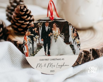 Op de afbeelding: Ronde houten kerstversiering met een trouwfoto van een stel. Het heeft een rood lint om op te hangen en de tekst "OUR FIRST CHRISTMAS AS Mr. & Mrs Longhorn 2023".