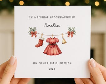 May include: A square white greeting card featuring the text TO A SPECIAL GRANDDAUGHTER, Amelia, and ON YOUR FIRST CHRISTMAS 2025. The design shows a clothesline with a red stocking, a red dress, and a gold bell, accented by green leaves and red bows.