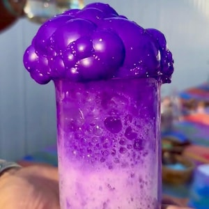 Puede incluir: Un vaso de precipitados de vidrio lleno de un líquido morado y cubierto con una gran nube de espuma morada.