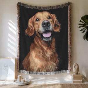 Peut inclure: Une couverture tissée avec un portrait de chien Golden Retriever sur fond noir. Le chien a un pelage brun clair, la gueule ouverte et la langue rose. La couverture a une bordure à franges.