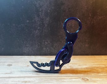 Könnte beinhalten: Ein dunkelviolettes, 3D-gedrucktes Klappmesser-Spielzeug im Karambit-Stil mit einem kreisförmigen Fingerring am Griffende, einer gebogenen, gezackten Klinge und mehreren beweglichen Gelenken, die durch kleine Schrauben miteinander verbunden sind.