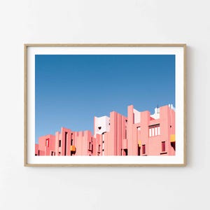 Può includere: Stampa incorniciata con una vivace fotografia architettonica. L'immagine mostra una serie di edifici rosa contro un cielo blu chiaro e luminoso. Gli edifici hanno un design geometrico unico con varie forme e dimensioni.