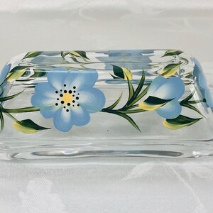 May include: A clear glass butter dish with a blue and yellow floral design. The flowers are painted on the glass and have a delicate, hand-painted look.