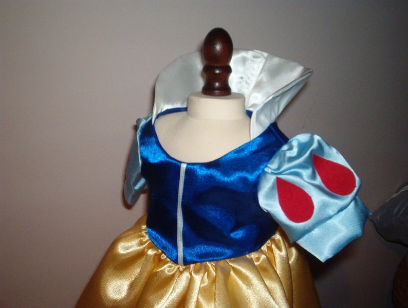 K&ouml;nnte beinhalten: Ein blaues und wei&szlig;es Satin-Kleid mit roten Akzenten. Das Kleid hat einen wei&szlig;en Kragen und Puff&auml;rmel mit roten Tropfenformen. Der Rock ist aus gelbem Satin.