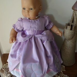May include: A doll wearing a lavender dress with a white underskirt and a pink ribbon. The dress has a floral design and the doll is wearing a pearl necklace.