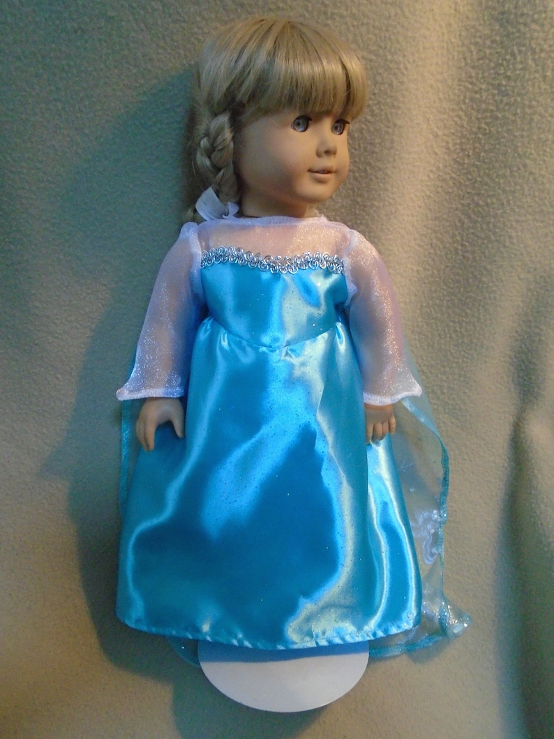 Peut inclure: Une poup&eacute;e portant une robe en satin bleu clair avec une bordure en dentelle blanche et une cape bleue transparente. La robe a une texture scintillante.