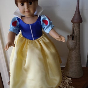 K&ouml;nnte beinhalten: Eine Puppe, die als Schneewittchen verkleidet ist, tr&auml;gt ein blaues und gelbes Kleid mit einem wei&szlig;en Kragen und roten Akzenten. Die Puppe hat braunes Haar und braune Augen. Die Puppe steht auf einer wei&szlig;en H&auml;kelspitze neben einem braunen Keramik-Schloss.