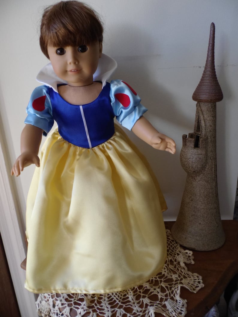 K&ouml;nnte beinhalten: Eine Puppe, die als Schneewittchen verkleidet ist, tr&auml;gt ein blaues und gelbes Kleid mit einem wei&szlig;en Kragen und roten Akzenten. Die Puppe hat braunes Haar und braune Augen. Die Puppe steht auf einer wei&szlig;en H&auml;kelspitze neben einem braunen Keramik-Schloss.