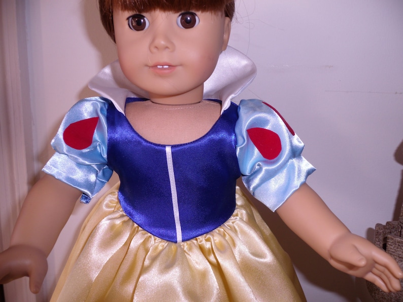K&ouml;nnte beinhalten: Eine Puppe, die ein blaues und wei&szlig;es Kleid mit roten Akzenten tr&auml;gt. Das Kleid hat einen wei&szlig;en Kragen und Puff&auml;rmel. Die Puppe hat braunes Haar und blaue Augen.