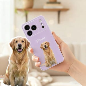 Op de afbeelding: Een lila telefoonhoesje met een golden retriever hond en de naam "Peanut" in script. De hoes heeft een camera-uitsparing en wordt door een persoon vastgehouden. Een golden retriever hond staat ook op de achtergrond.