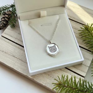 May include: A silver necklace with a pendant featuring a detailed cat face design. The octagonal pendant is presented in a white box. The necklace is displayed on a wooden surface with pine needles and a pine cone.