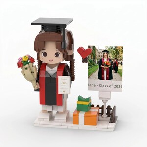 May include: A graduation-themed Lego figure of a person in a cap and gown, holding a bouquet and a certificate. A photo of a graduate is displayed next to the figure. The figure is standing on a white base with a gift box.
