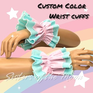 May include: Two pairs of custom color wrist cuffs in a light blue and pink color scheme. The cuffs are made of fabric and have a ruffled design. The text "CUSTOM COLOR WRIST CUFFS" is displayed at the top of the image.