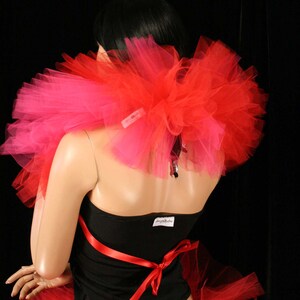 May include: A red and pink tulle tutu skirt with a black top and a red ribbon. The skirt is layered and has a fluffy texture. The top is a simple black tank top with a red ribbon tied around the waist. The model is wearing the skirt and top and is standing in front of a black background.