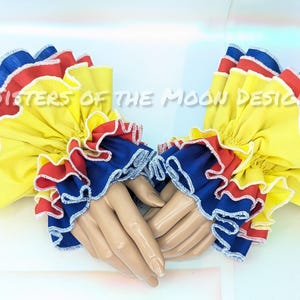 May include: A pair of ruffled wrist cuffs in yellow, red, and blue, with white trim. The cuffs are displayed on a mannequin hand. The text "Sisters of the Moon Design" is visible in the background.