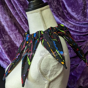 May include: A black fabric collar with colorful paint splatter design. The collar has a gathered elastic band and six points. The text "Sisters of the Moon Design" is visible on the collar.