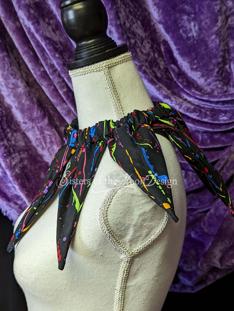 May include: A black fabric collar with colorful paint splatter design. The collar has a gathered elastic band and six points. The text "Sisters of the Moon Design" is visible on the collar.