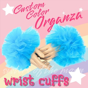 May include: Two light blue organza wrist cuffs. The cuffs are ruffled and voluminous, with a soft, airy texture. The image includes the text "Custom Color Organza" and "wrist cuffs" in pink, along with a rainbow background.