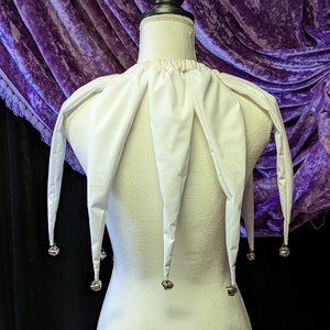 May include: White jester collar with five points and silver bells hanging from each point. The collar is on a white mannequin against a purple background.