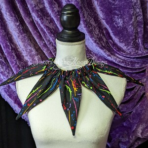 May include: A black fabric collar with a multi-colored paint splatter design. The collar has six points and is on a white mannequin. The text "Sisters of the Moon Design" is visible on the collar.