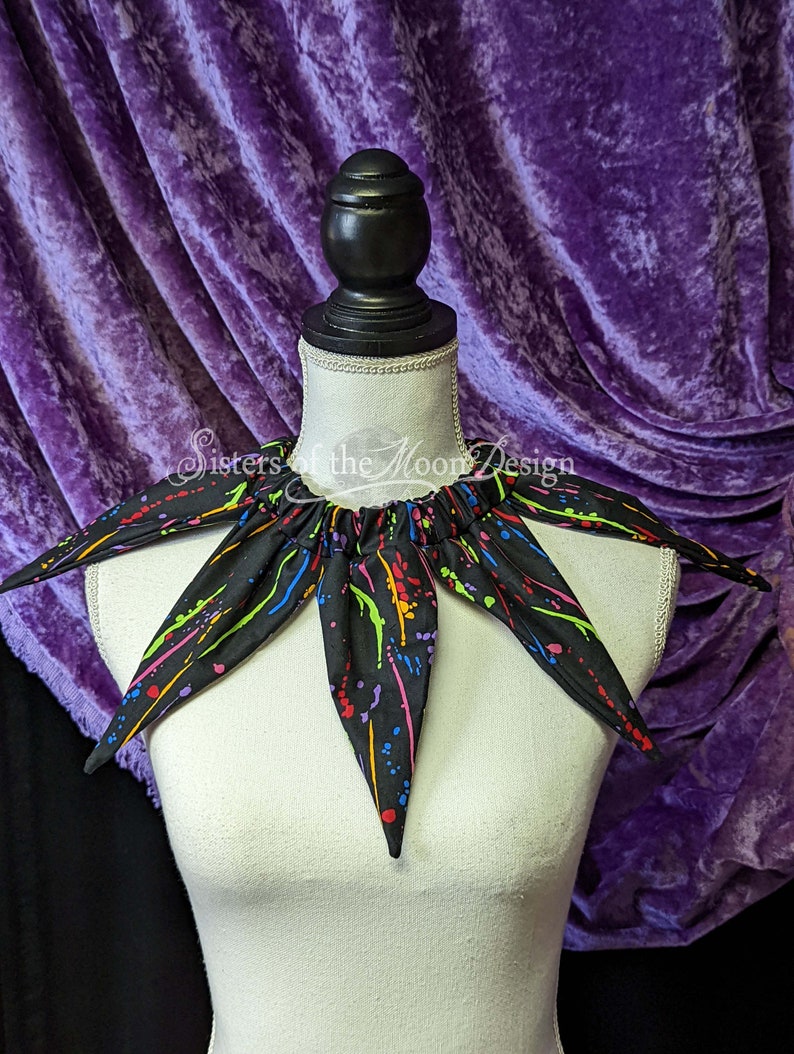 May include: A black fabric collar with a multi-colored paint splatter design. The collar has six points and is on a white mannequin. The text "Sisters of the Moon Design" is visible on the collar.