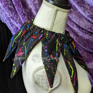 May include: A black fabric collar with colorful paint splatter design. The collar has six points and is made of a soft, stretchy fabric. The collar is on a white mannequin.