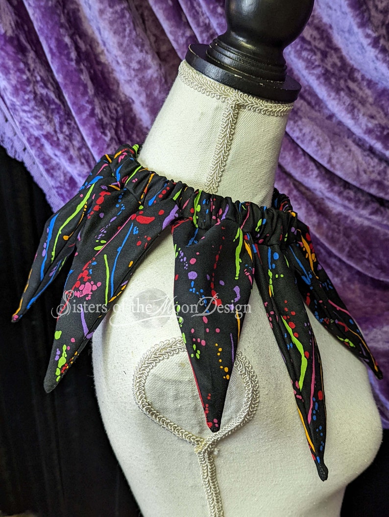 May include: A black fabric collar with colorful paint splatter design. The collar has six points and is made of a soft, stretchy fabric. The collar is on a white mannequin.