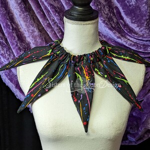 May include: Black fabric collar with a splatter paint design in rainbow colors. The collar has six points and is gathered at the back. The text "Sisters of the Moon Design" is visible on the collar.