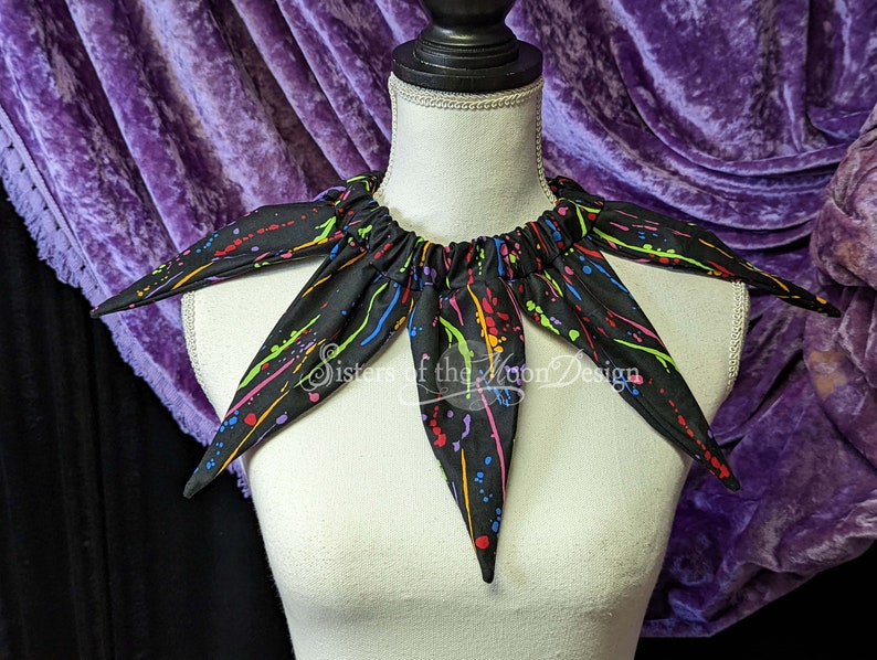 May include: Black fabric collar with a splatter paint design in rainbow colors. The collar has six points and is gathered at the back. The text "Sisters of the Moon Design" is visible on the collar.