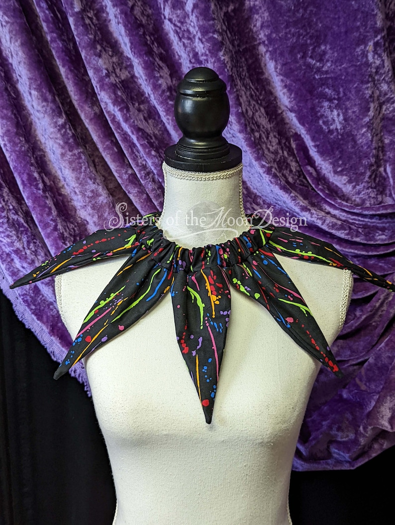 May include: A black fabric collar with a splatter paint design in rainbow colors. The collar is on a white mannequin. The text "Sisters of the Moon Design" is visible on the collar.