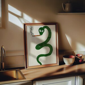 以下が含まれることがあります： 口にタバコを持った緑色のヘビの絵が額装されています。ヘビはリラックスした姿勢で丸まっています。絵は白い背景に描かれており、木製の額縁が付いています。