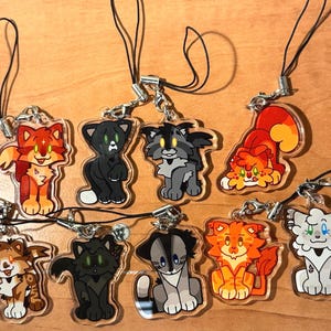 May include: A collection of nine cat-themed acrylic charms with black lanyards. The charms feature various cartoon cat designs in orange, black, gray, and white. Each charm has a silver-colored clasp.