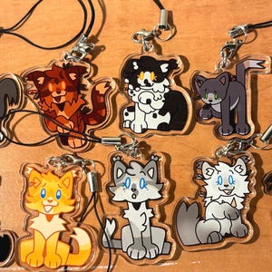 May include: A collection of nine acrylic cat charms with various designs. Each charm features a different cartoon cat in various colors, including gray, orange, black, and white. The charms are attached to black cords with silver-colored clasps.