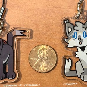 May include: Two clear acrylic cat charms with cartoon illustrations. One is a gray cat with green eyes and a red bow, the other is a gray and white cat with blue eyes. A penny is between them.