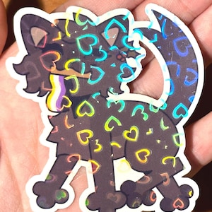 Pu&ograve; includere: Un adesivo con un design stilizzato di un gatto. Il gatto &egrave; marrone scuro con un motivo di cuori colorati. L'adesivo ha un effetto olografico, creando un aspetto scintillante. Il gatto ha un collare arcobaleno.