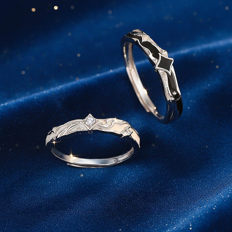 K&ouml;nnte beinhalten: Zwei silberne Ringe mit aufwendigen Designs. Ein Ring hat ein schwarzes Emailleband mit einem rautenf&ouml;rmigen Akzent, der andere ein wei&szlig;es Design mit einem zentralen Diamanten. Die Ringe sind vor einem dunkelblauen Hintergrund platziert.
