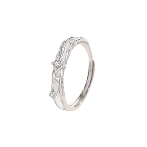 K&ouml;nnte beinhalten: Silberfarbener Ring mit einem dekorativen wei&szlig;en Design und zwei kleinen, klaren Edelsteinen. Der Ring hat ein offenes Design und ist wahrscheinlich verstellbar. Das dekorative Design hat ein flie&szlig;endes, organisches Muster.